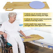 Older woman seated in a wheelchair beside a bed, with a wooden transfer board bridging the two surfaces and a blue rotating seat disc positioned near the wheelchair.